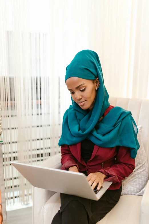 a woman wearing a hijab sits in a chair using her laptop