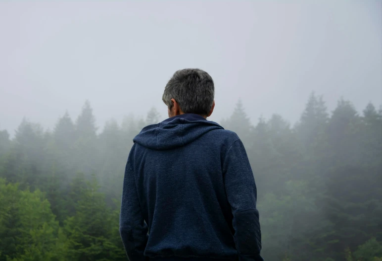 the back view of a man in a hoodie standing on a hill overlooking fog covered trees