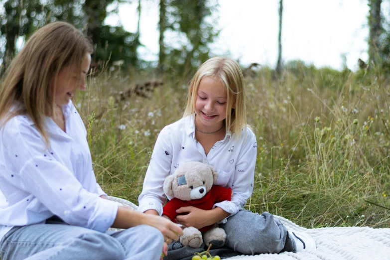 two girls sit in the grass and laugh while holding stuffed animals