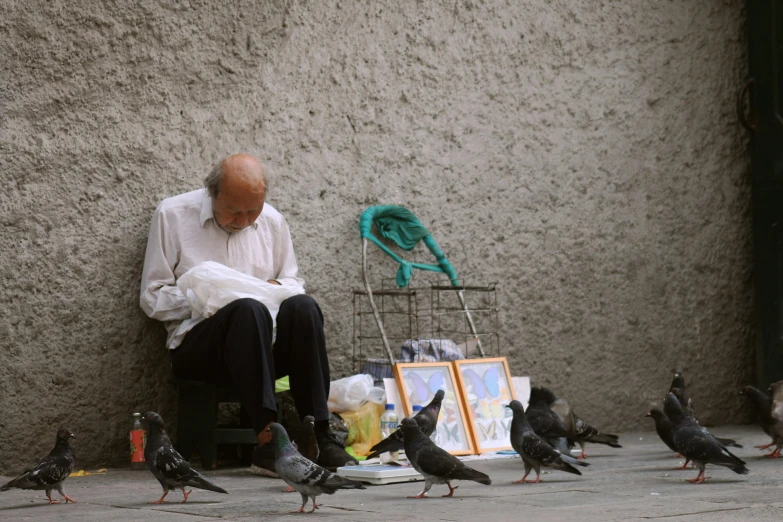 the man is sitting against the wall surrounded by many pigeons