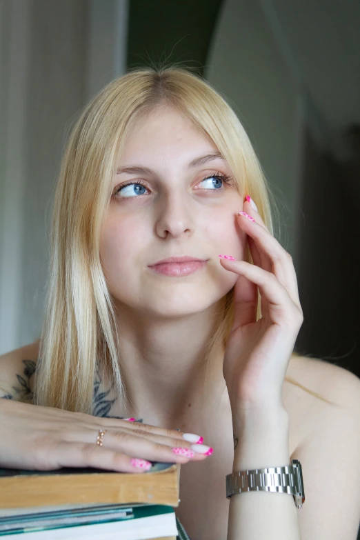 a young blonde with tattoos and pink fingernails poses in front of stacks of books