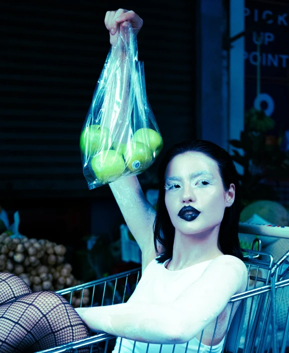 a woman with black makeup and black eye makeup sits in an empty shopping cart holding a grocery bag with two green apples in it