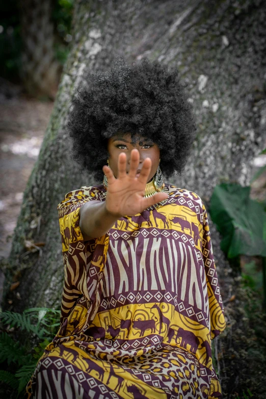 a young woman in a ze print dress stands next to a tree while holding her hand up