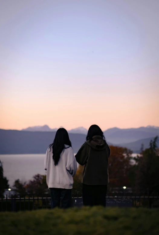 two young women in coats stand back to back on a hillside overlooking water and mountains