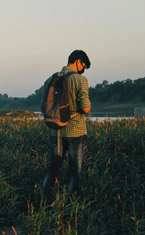 man in plaid shirt and jeans in tall grass field with book bag on his back