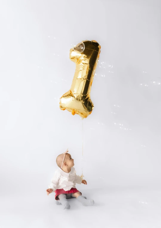 a baby in a red shorts sits under a gold number one balloon