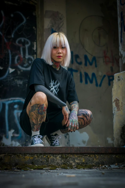 tattooed girl wearing black t - shirt squatting down on a sidewalk