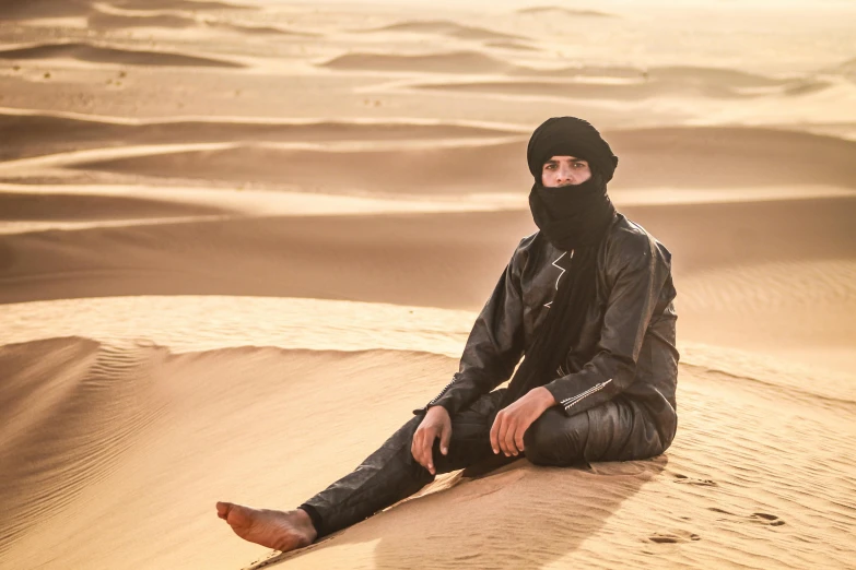 there is a man in a black hat and black jacket sitting in the desert
