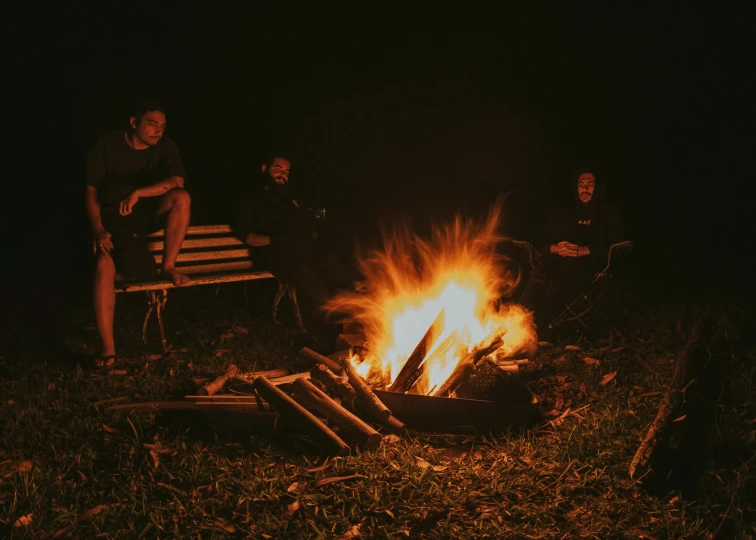 three people sitting around a campfire with a bench and a fire in the background