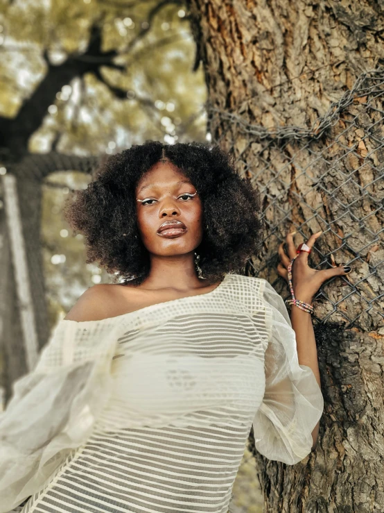 a woman wearing white poses with her hands on the side of a tree