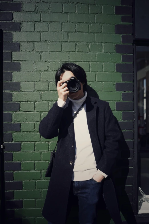 man in black coat and glasses standing against a brick wall