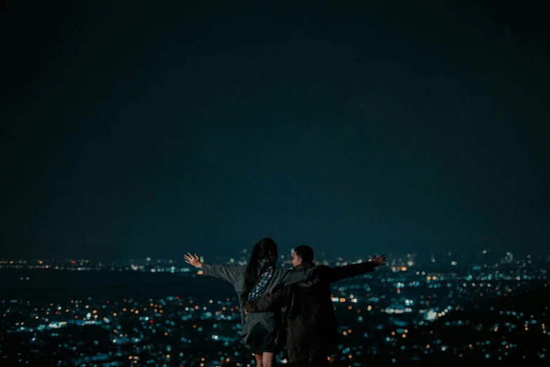 a man and woman stand on a mountain while holding hands out to the city lights