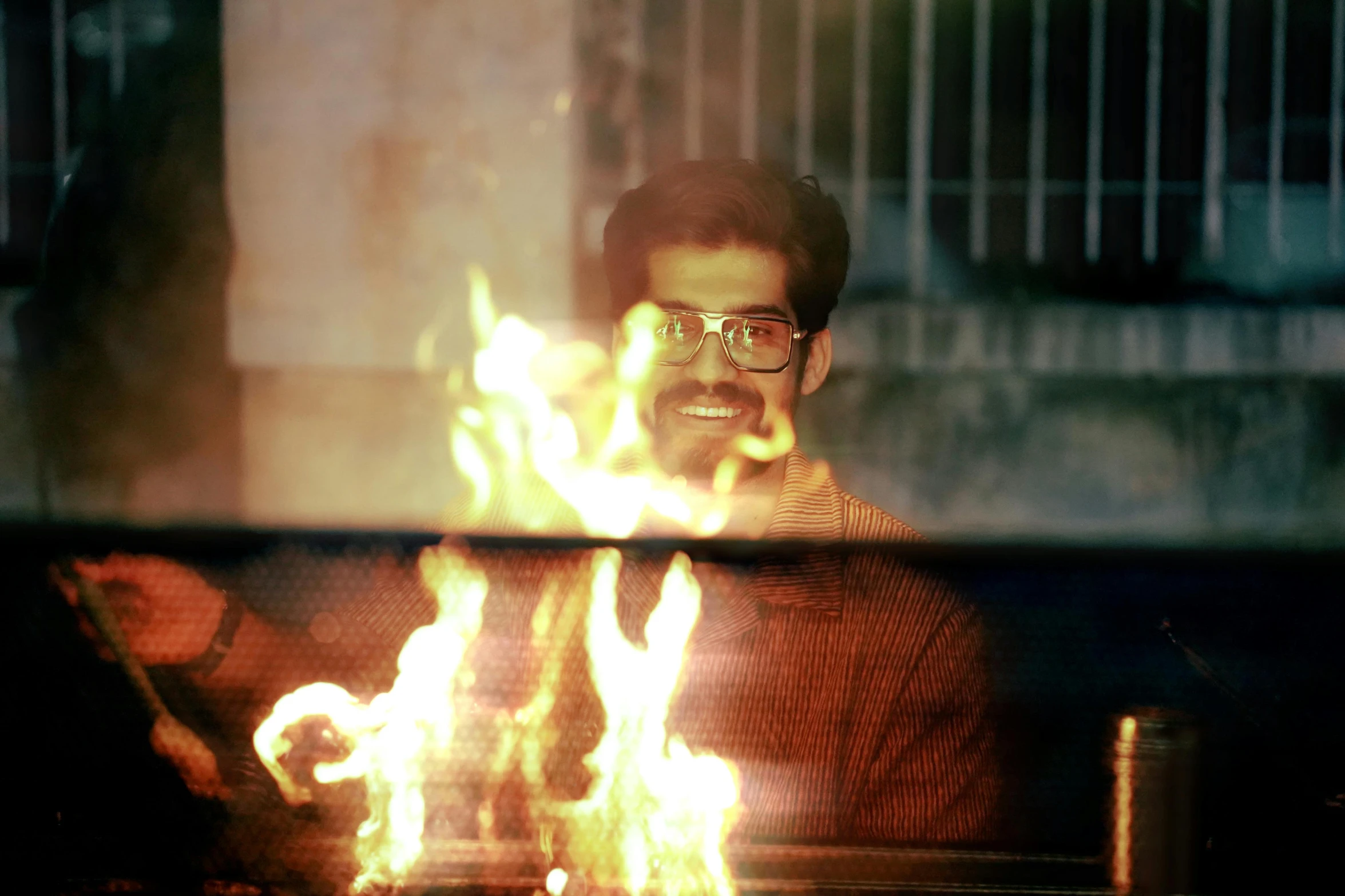 man with glasses sitting on a bench in front of a large fire