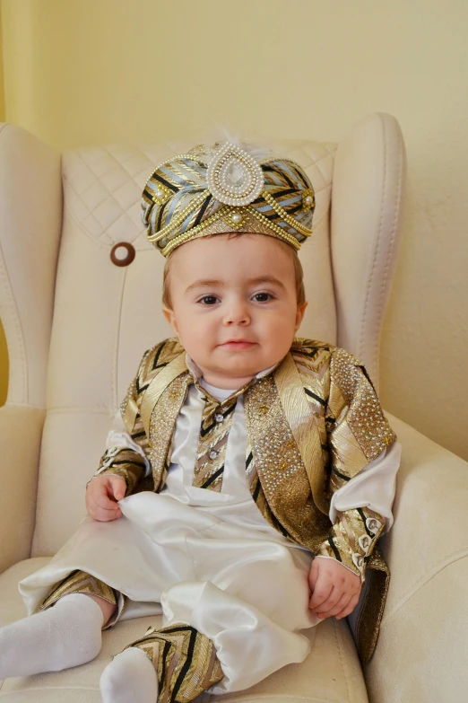 a baby sitting in a white chair wearing a gold colored suit