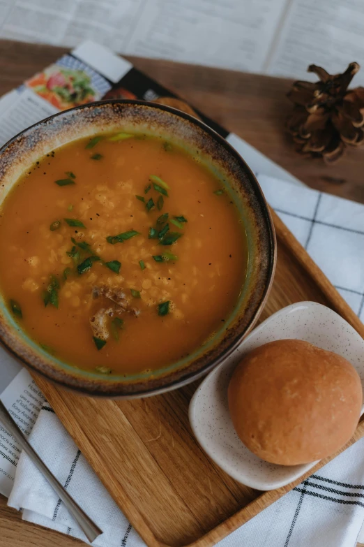 a bowl of soup next to a roll on top of a wooden tray