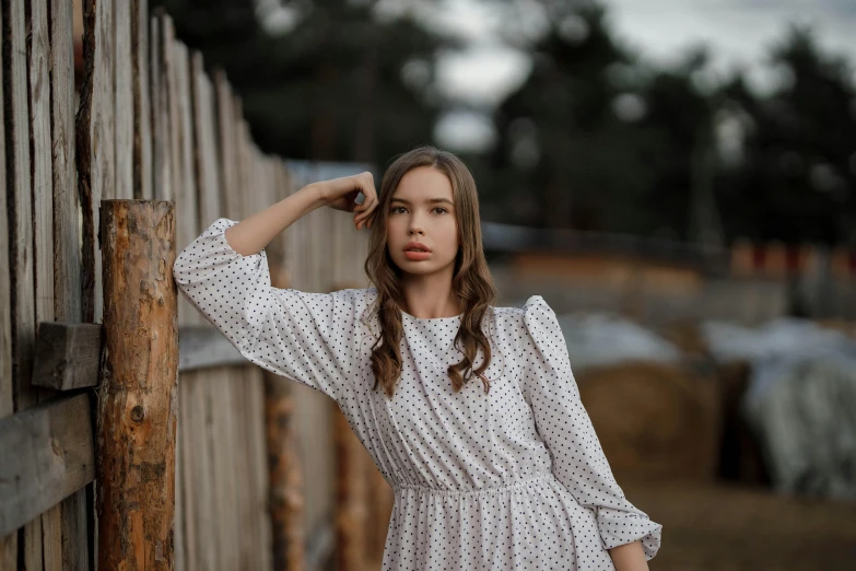 an attractive young woman in a dress leans against a fence
