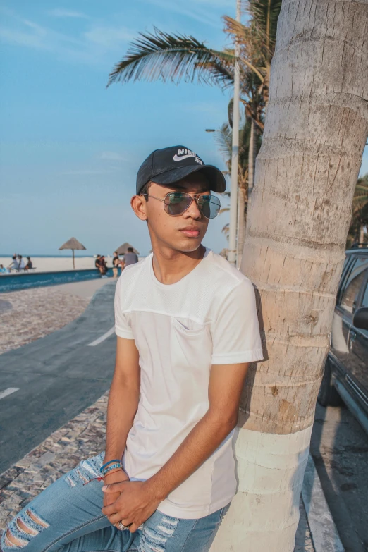 a man in a white shirt and sunglasses leaning against a tree on the beach