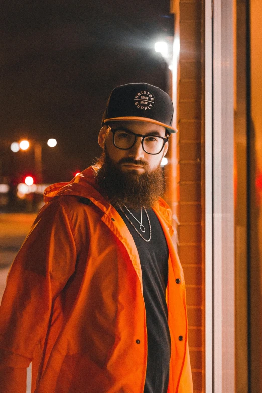 a bearded man with glasses wearing an orange jacket, black hat and dark glasses