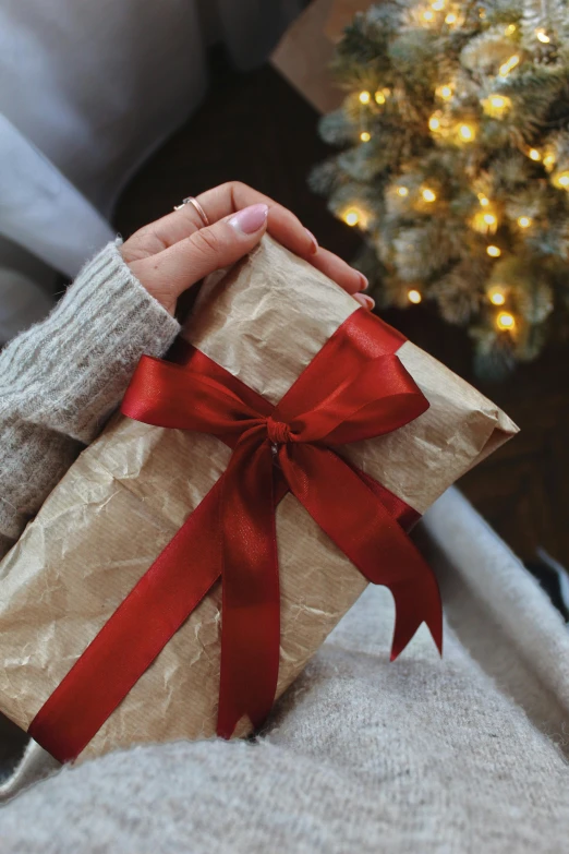 a small present wrapped in brown paper with a red ribbon and bow on it, sitting on the side of a christmas tree