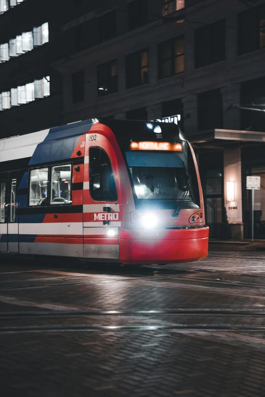 a red, white and blue train traveling down a street next to tall buildings