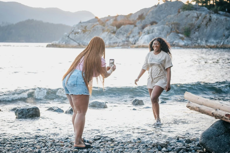 two women are on the beach one is holding a drink and one is standing in the water