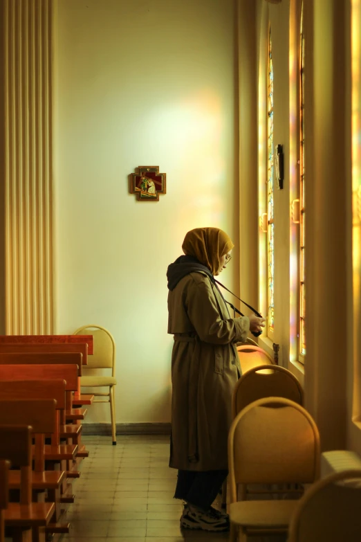 a woman with a hair covering standing next to an empty chair in front of stained glass windows