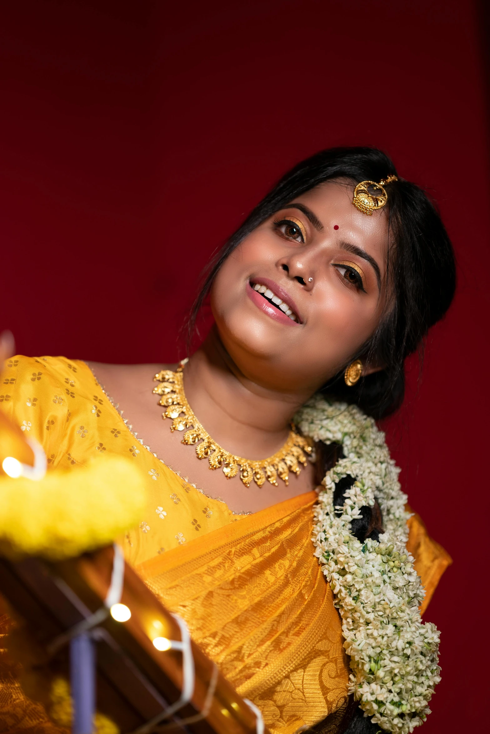 a girl in an indian outfit and garland stands near a lit lamp