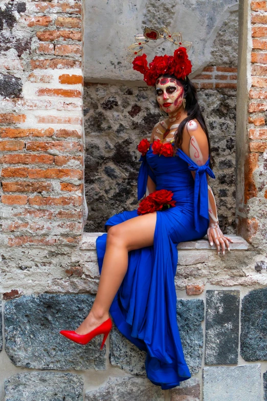 a woman wearing an elaborately decorated face and blue dress sitting on a stone ledge