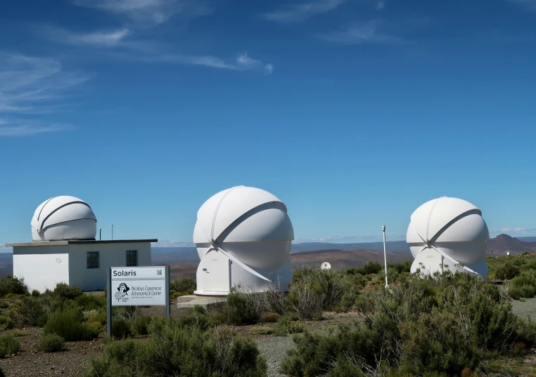 three large white round structures stand in front of scrubby land