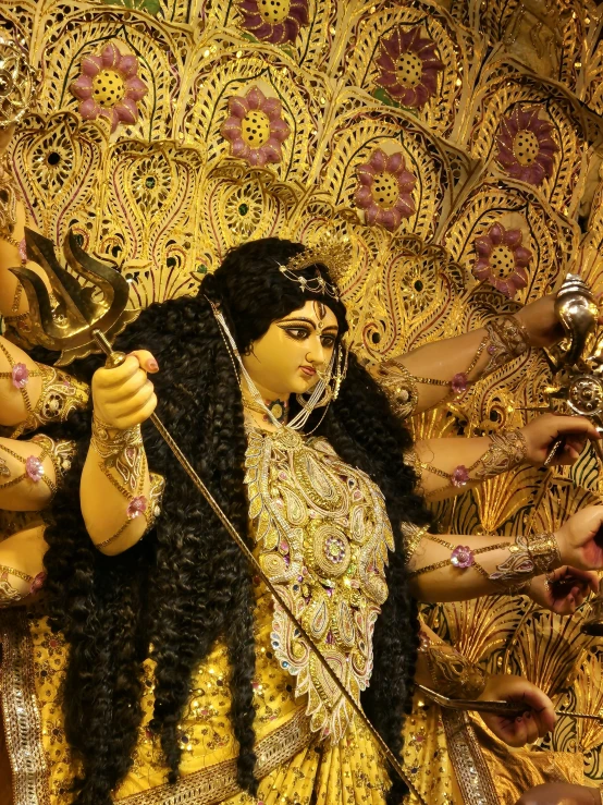 woman with long hair and gold clothing holds a staff and lights herself in front of an ornately designed wall