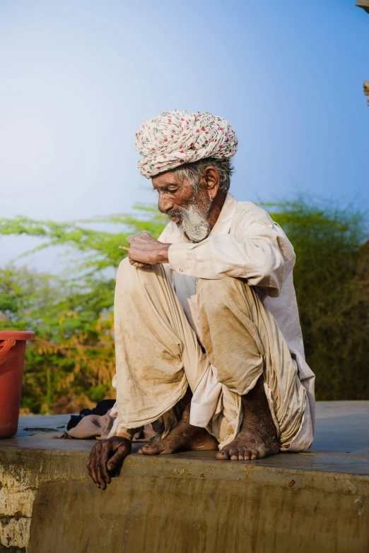 an old man with a white turban and floral head cover, sits on the ledge of a building