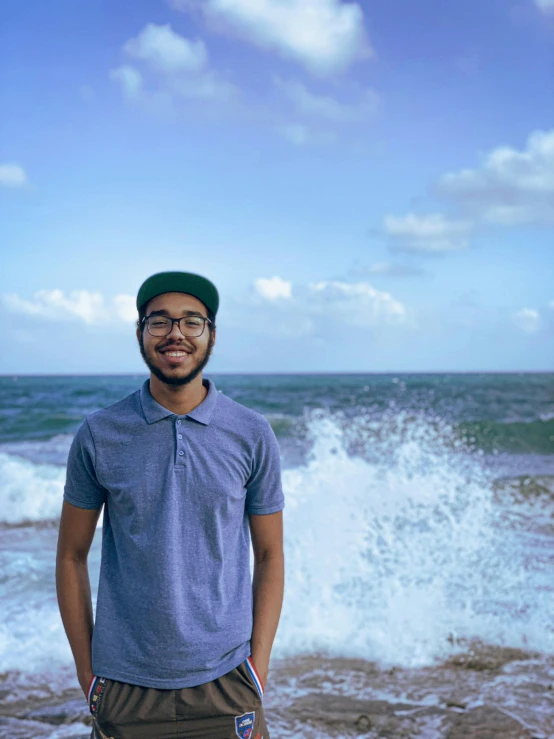 a young man with glasses stands near the ocean shore