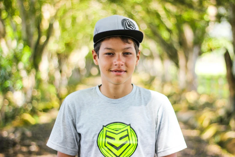 a boy in grey shirt and black hat standing in front of trees