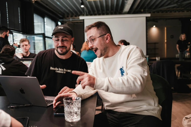 a guy laughing at someone on his laptop in front of a guy with a glass of water