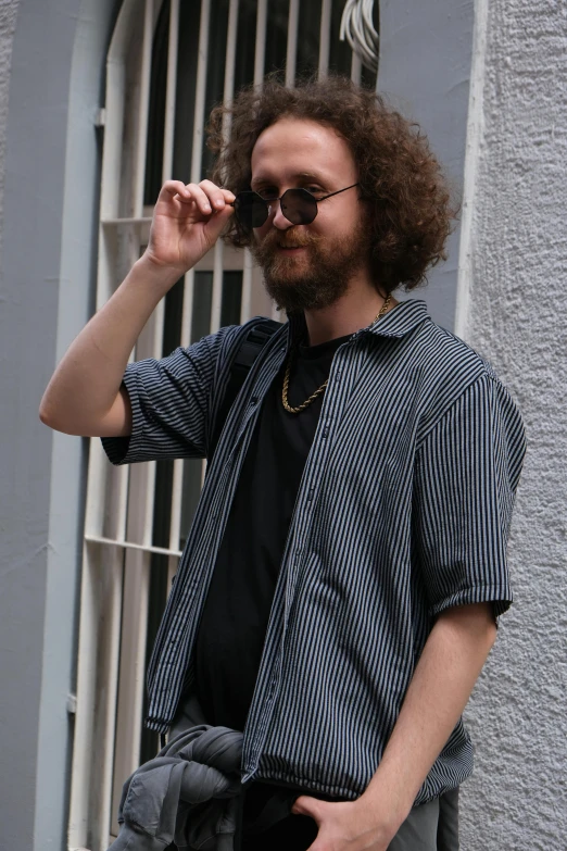 a man wearing glasses, a blue shirt, and grey pants standing in front of a grey building