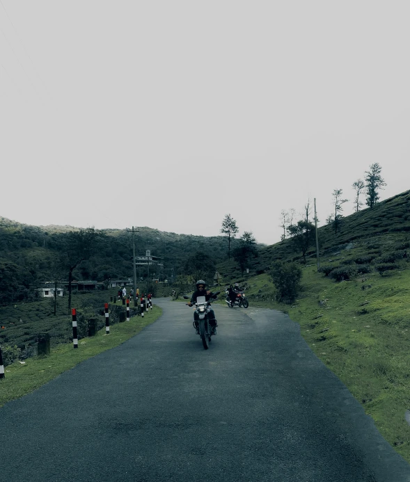 two people on motorcycles riding down a road while others stand around and watch