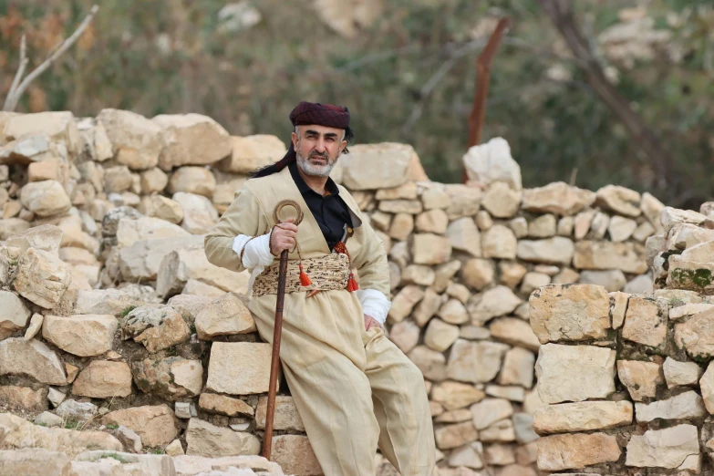 a man with a beard and long brown hair, dressed in an outfit, standing in a stone wall