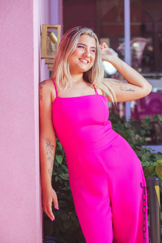 a woman with long blonde hair is leaning on the pink wall and looking at the camera