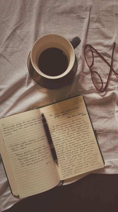 the book is opened on a bed with glasses, cup and pen