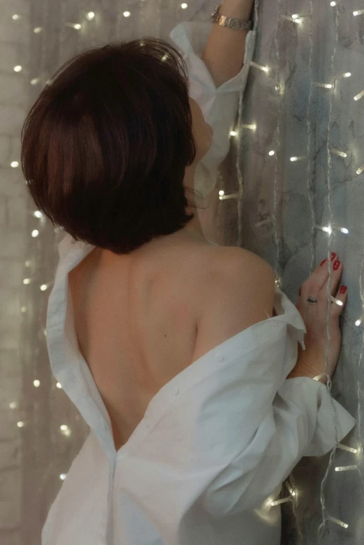 a woman with dark hair wearing a white shirt looks through a window at light - covered curtains