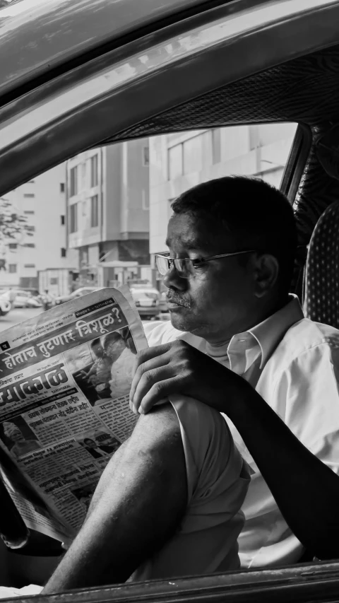 an asian man sitting in the driver's seat of a car reading a newspaper