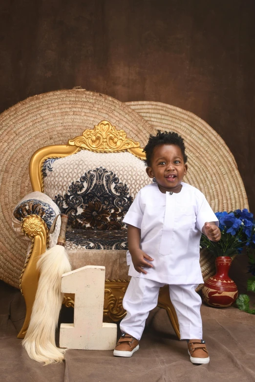 a little boy standing by a gold headdress and wooden rocking chair