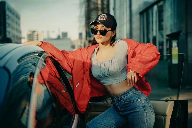 a woman in jeans and a baseball cap sitting on the hood of her car