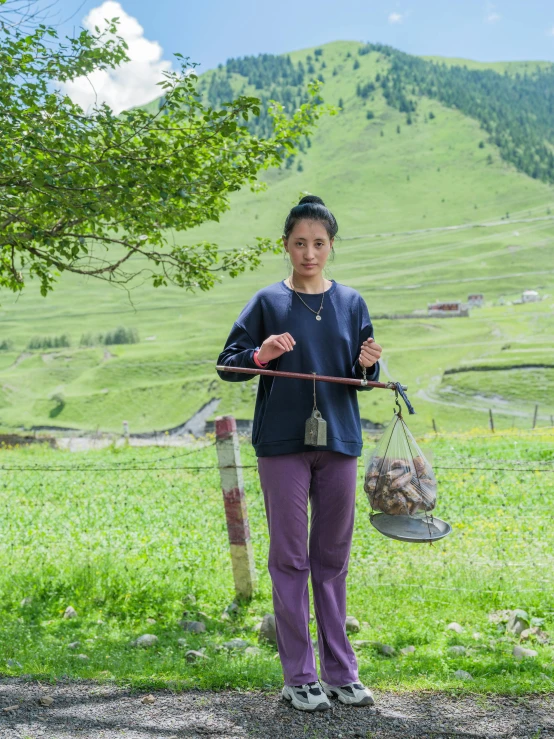 a young woman holding a large metal pot and bag while standing on a dirt road in the mountains