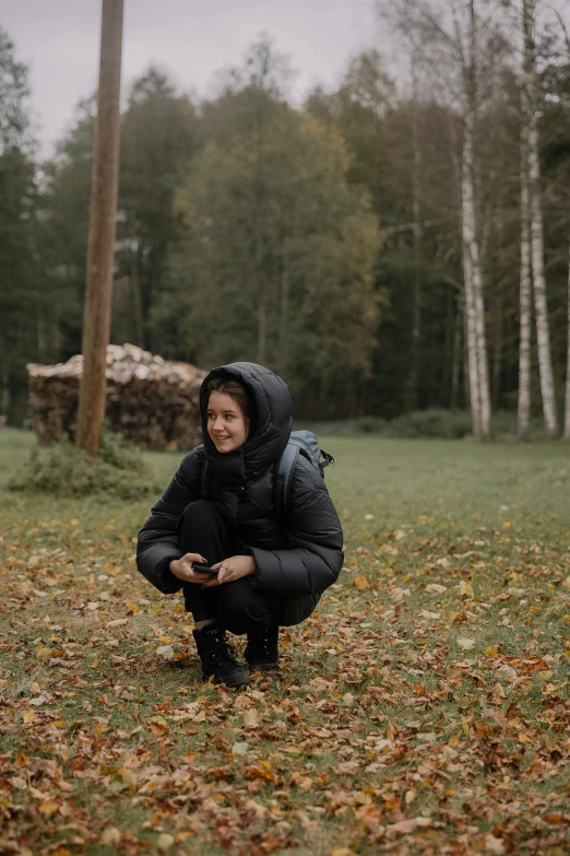 a woman in a jacket squatting on the grass with leaves all over her