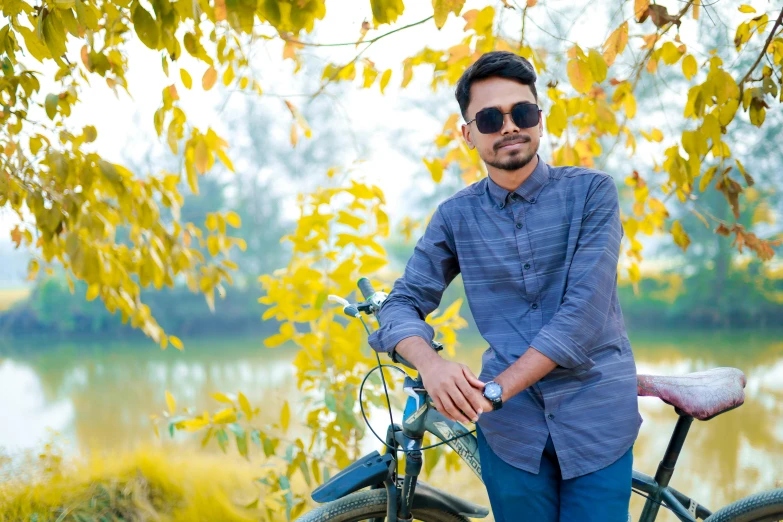 the man wearing sunglasses and a plaid shirt sits near his bicycle by a river