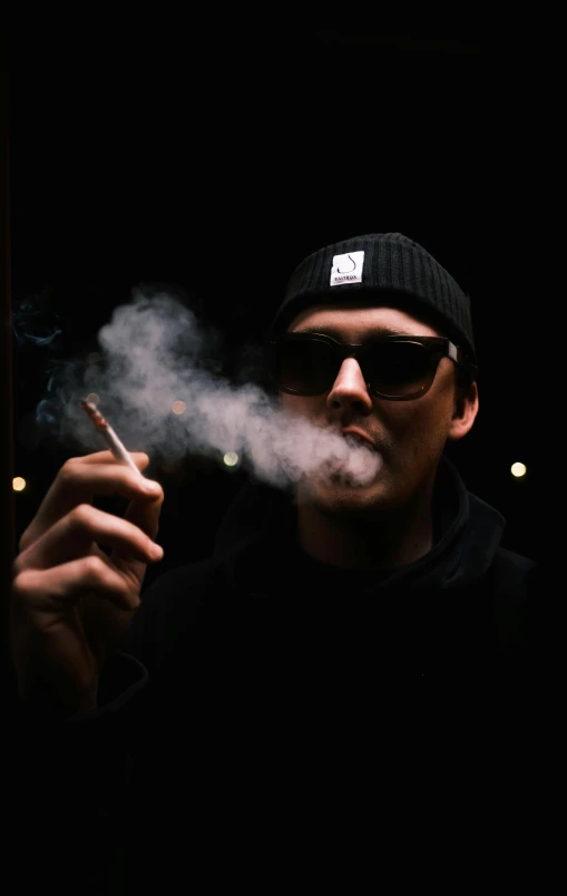 a man smokes on a dark surface while wearing a hat and sunglasses