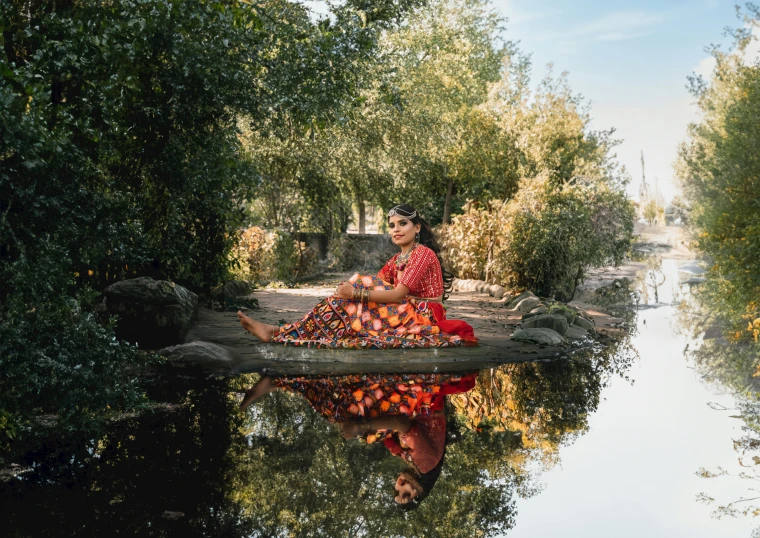 woman in red and green sitting on log on stream