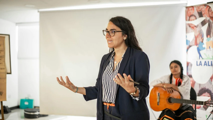 a woman in glasses standing and talking to someone with guitar instruments behind her