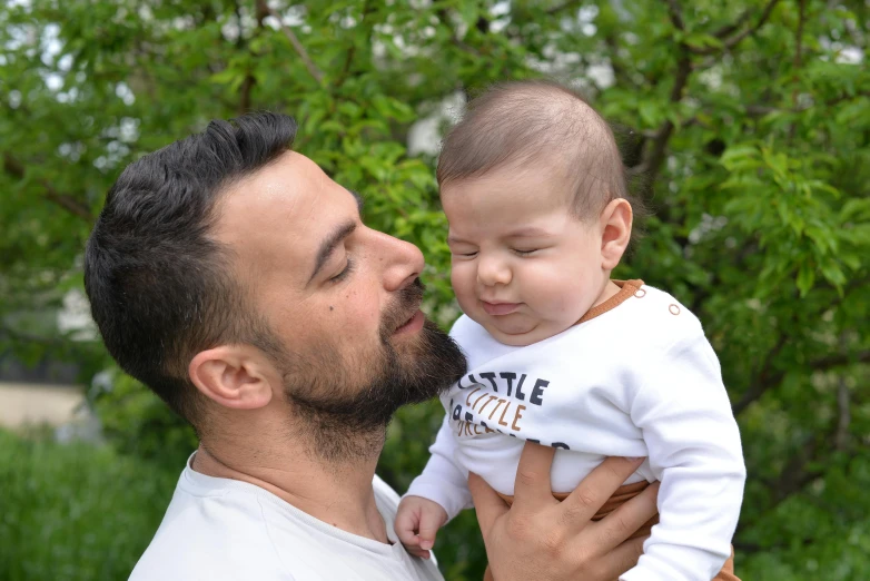 man with beard holding baby in yard near trees